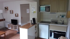 Een kleine keuken met een magnetron, spoelbak en een kleine koelkast. In de woonruimte staat een houten tafel en een televisie.