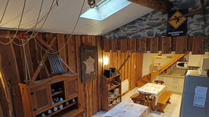 Une cuisine rustique aux murs en bois, dotée d’une lucarne, d’un panneau d’avertissement signalant la présence de gaz dangereux et d’une table recouverte d’une nappe matelassée.