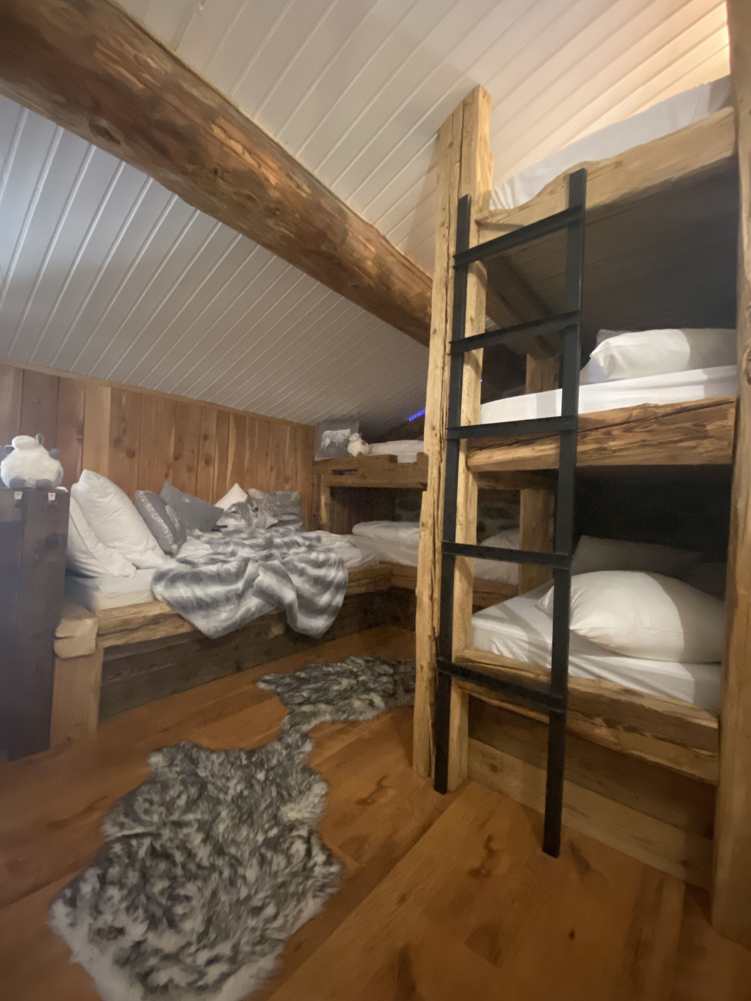 A cabin with wooden walls and a sloped ceiling. There are bunk beds with white bedding and a ladder. A fur rug is on the wooden floor.