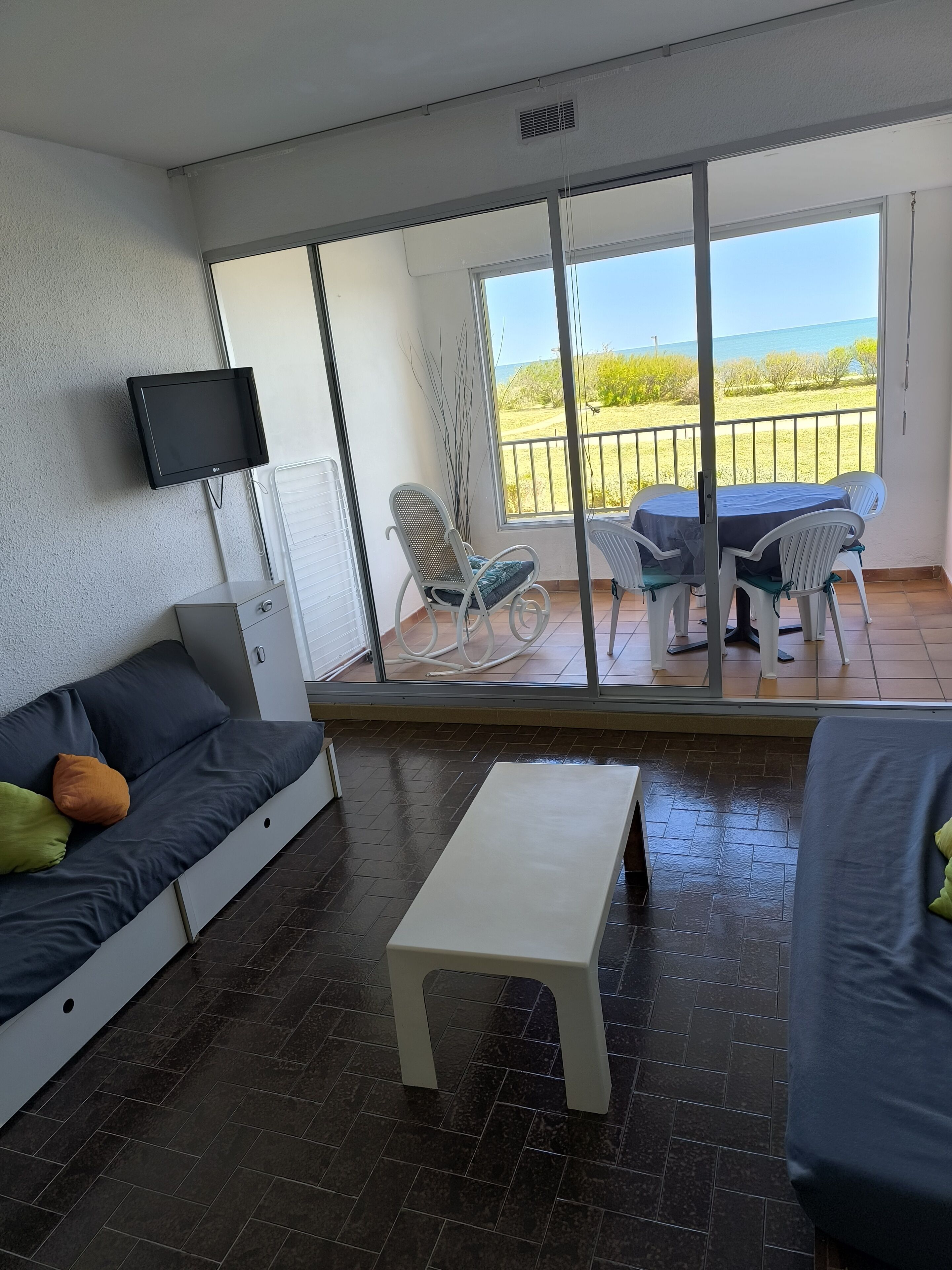 A modern living room with a sofa, a TV, a small table, and a balcony with a view of the sea.