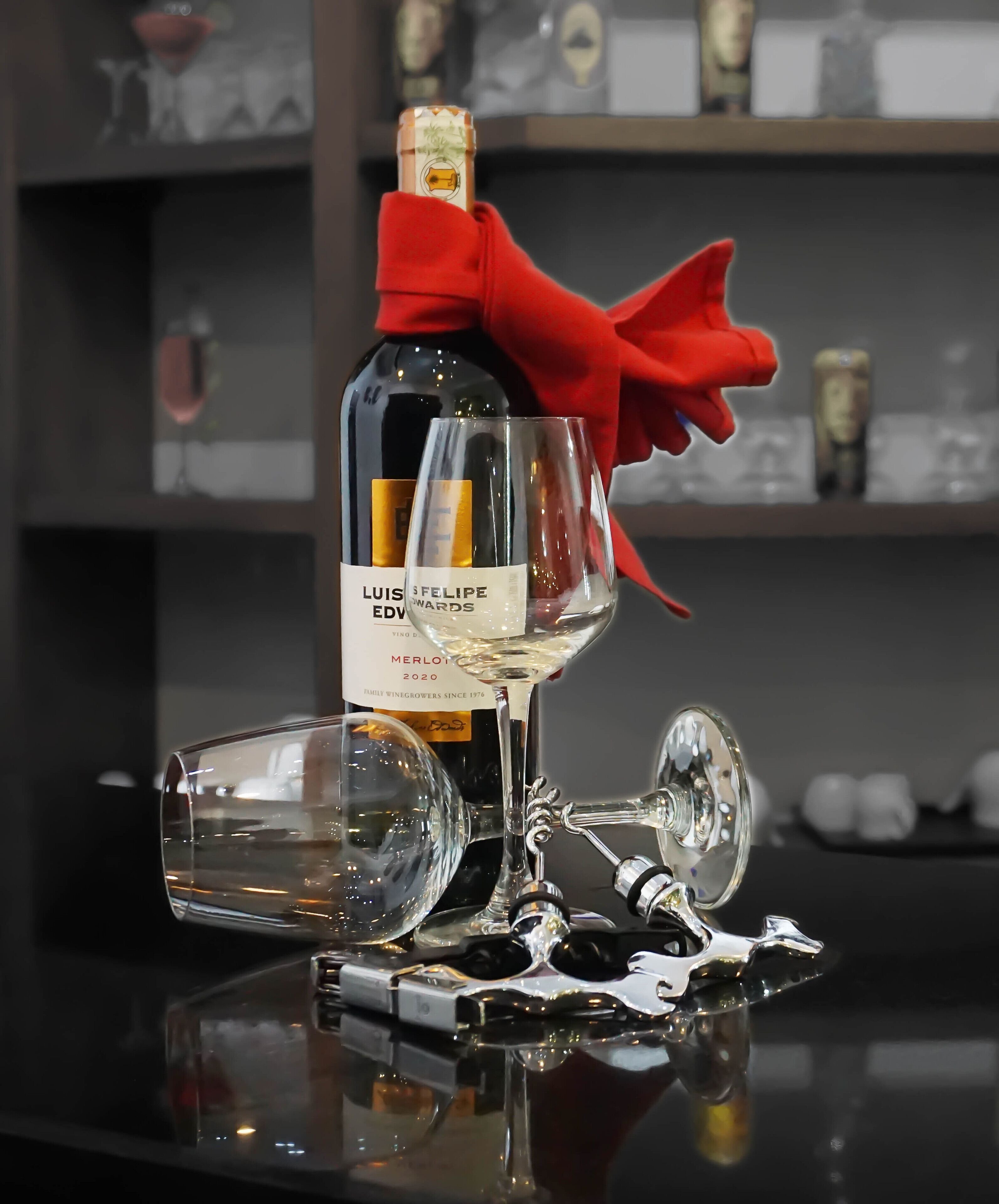 A bottle of Luis Eduardo Merlot 2020 wine with a red bow, a wine glass, and a decanter on a reflective surface.