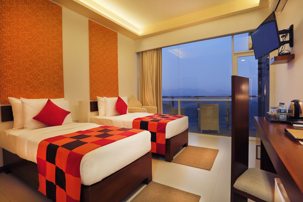 Habitación de hotel con dos camas, un escritorio, televisión y balcón con vistas.