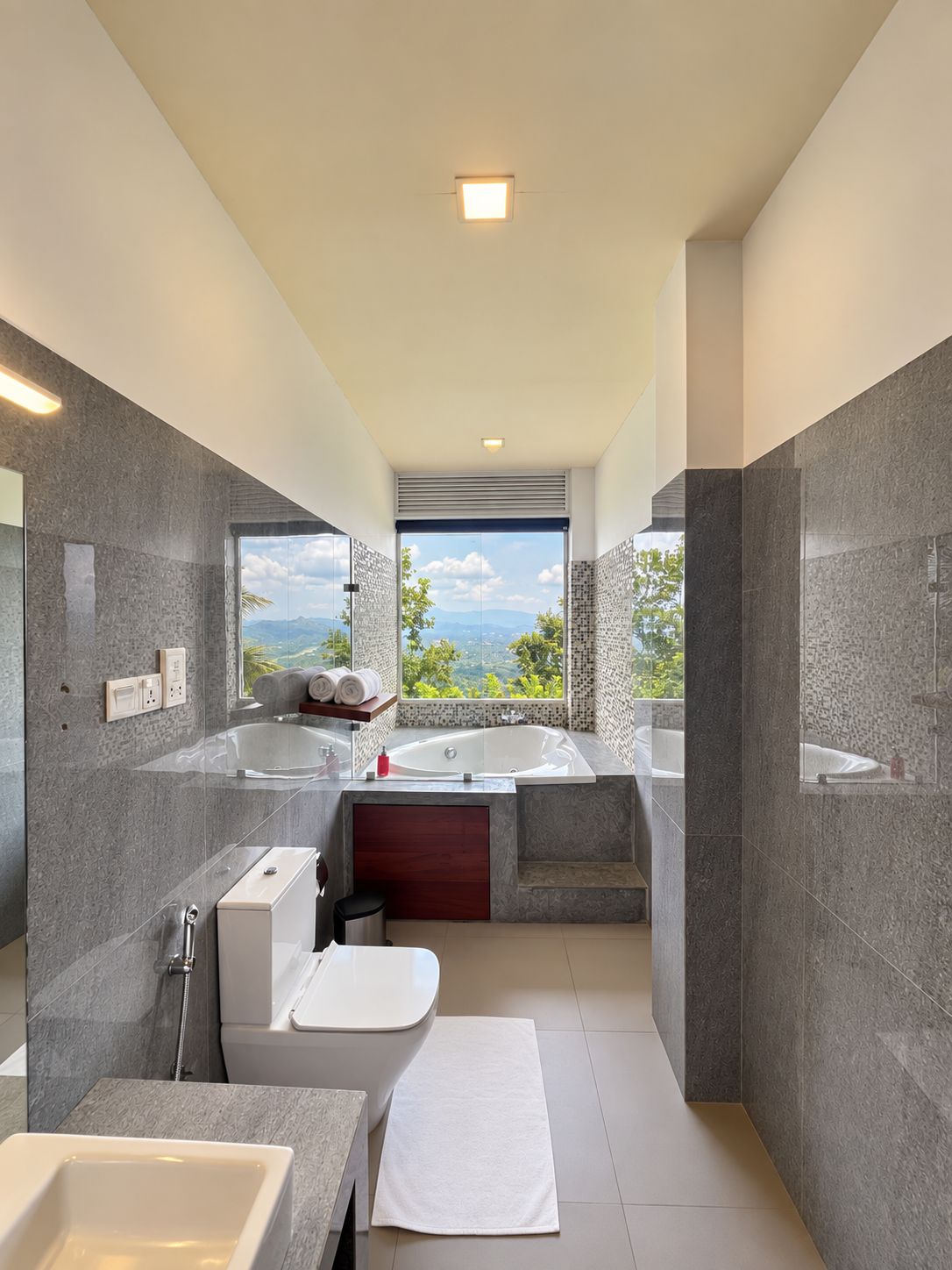 A modern bathroom with a bathtub, toilet, and sink, featuring a large window with a mountain view.