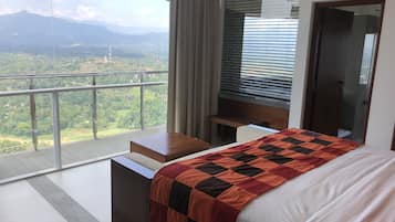 A bedroom with a large bed, a wooden nightstand, a desk, and a view of a mountainous landscape through large windows.