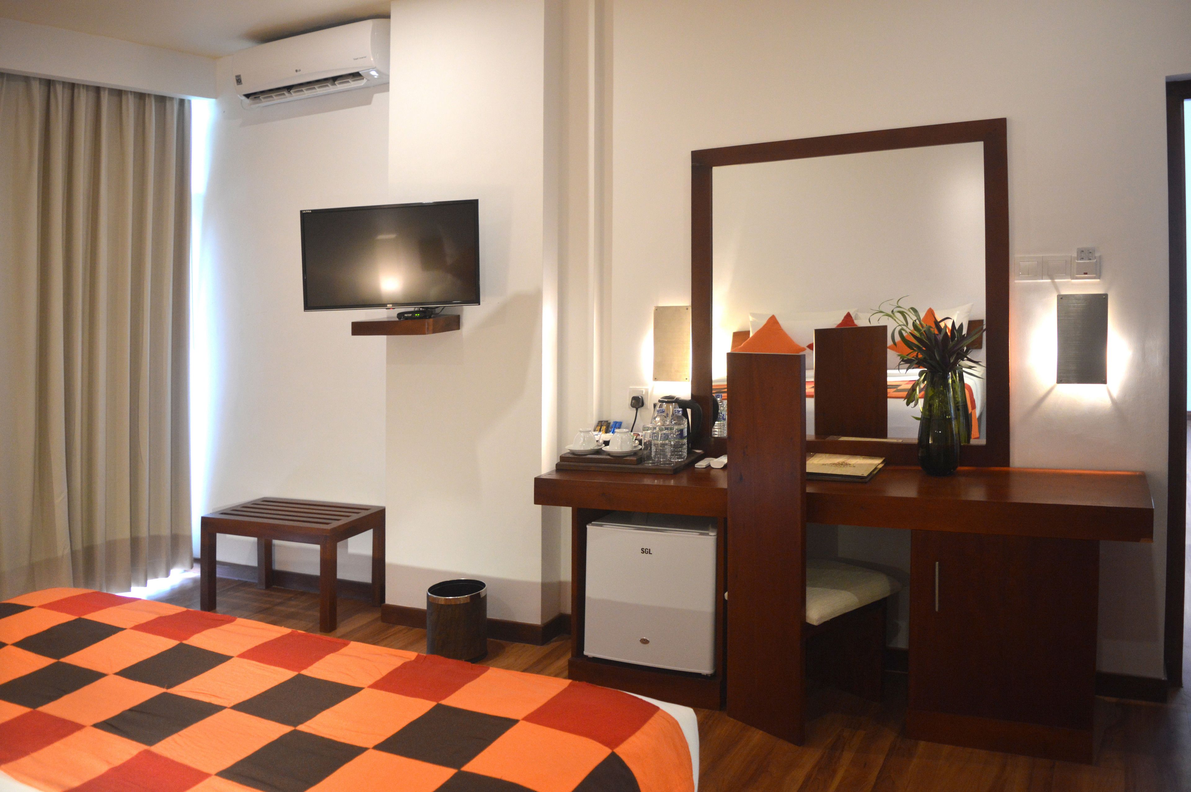 A hotel room with a bed, a desk with a mirror, a television, a small refrigerator, and a trash can.