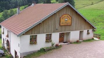 Un bâtiment de style rustique, rappelant une grange, avec un toit en pente, une enseigne en bois et un petit porche agrémenté de plantes en pot.