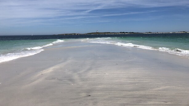 A wide sandy beach with gentle waves, a clear sky, and a distant shoreline with buildings and a lighthouse.