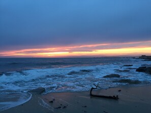 A beach with waves crashing onto the shore, a sunset in the background, and a piece of driftwood on the sand.