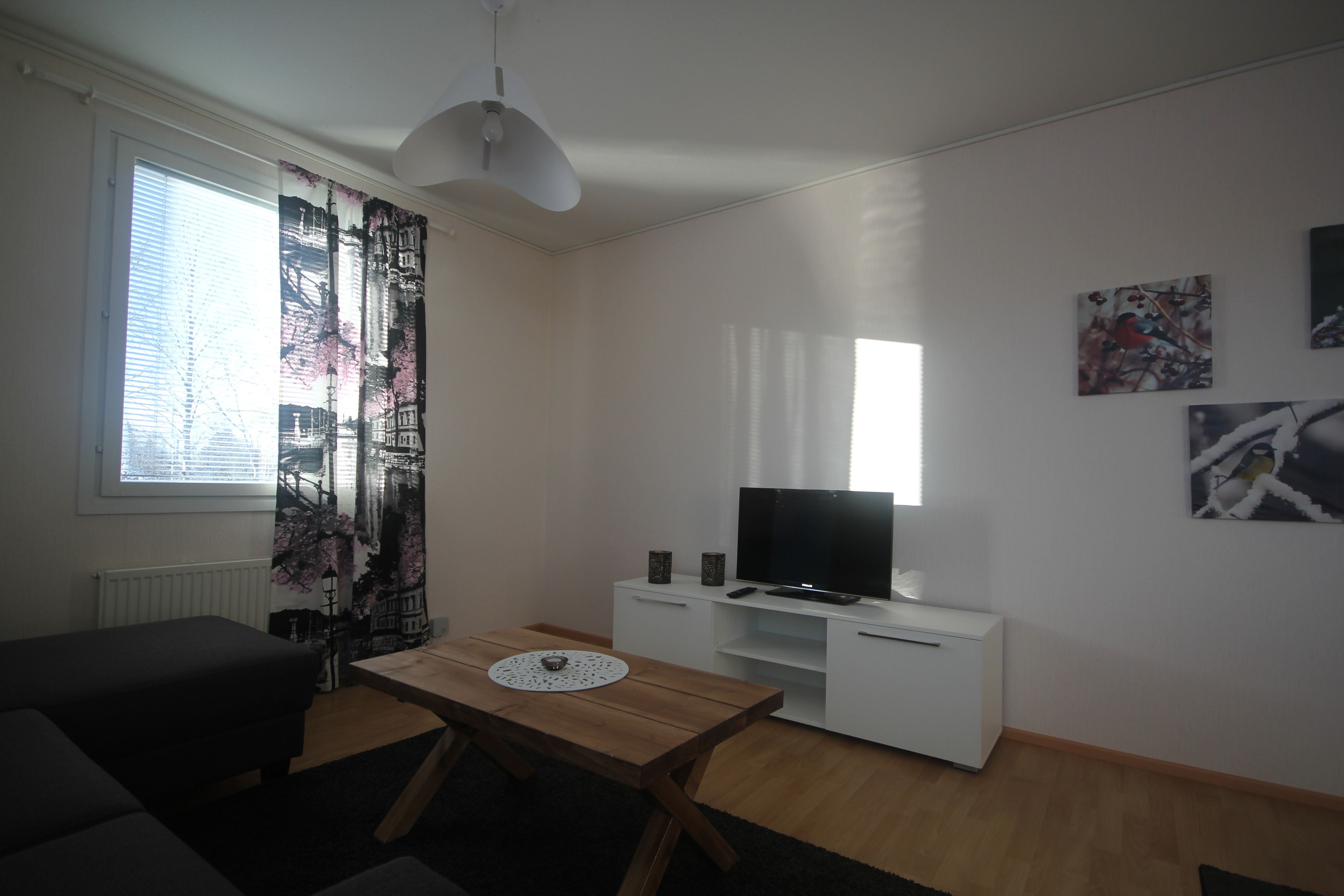 A living room with a sofa, a wooden table, a TV on a white cabinet, and a window with curtains.
