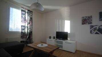 A living room with a sofa, a wooden table, a TV on a white cabinet, and a window with curtains.
