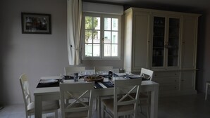 Ein Esszimmer mit Tisch für vier Personen, weißen Stühlen, einem Schrank mit Glastüren und einem fensterbepflanzten Fenster.