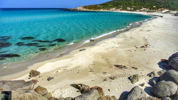 Uma praia com águas turquesa cristalinas, areal arenoso e afloramentos rochosos.