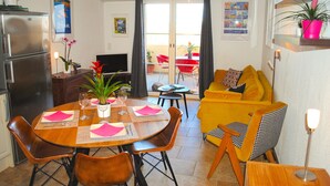 Uma área de refeições com uma mesa de madeira posta para quatro pessoas, cercada por cadeiras. Há também uma cozinha com geladeira e uma mesa de jantar com cadeiras. Um sofá e uma mesinha com abajur são visíveis ao fundo.