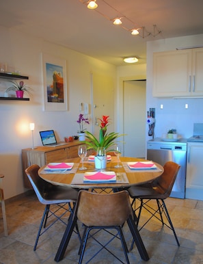 A dining area with a wooden table, four chairs, and a laptop on the side. There is a potted plant on the table and a framed picture on the wall.