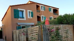 A two-story building with a wooden fence, a balcony with a red umbrella, and a gravel driveway. - Vacation home 2 bedrooms, inner courtyard, seaside, 5 places max WiFi parking (Torreilles)