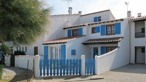 Un immeuble de deux étages avec une façade blanche et des volets bleus, une entrée fermée par un portail et un petit balcon.