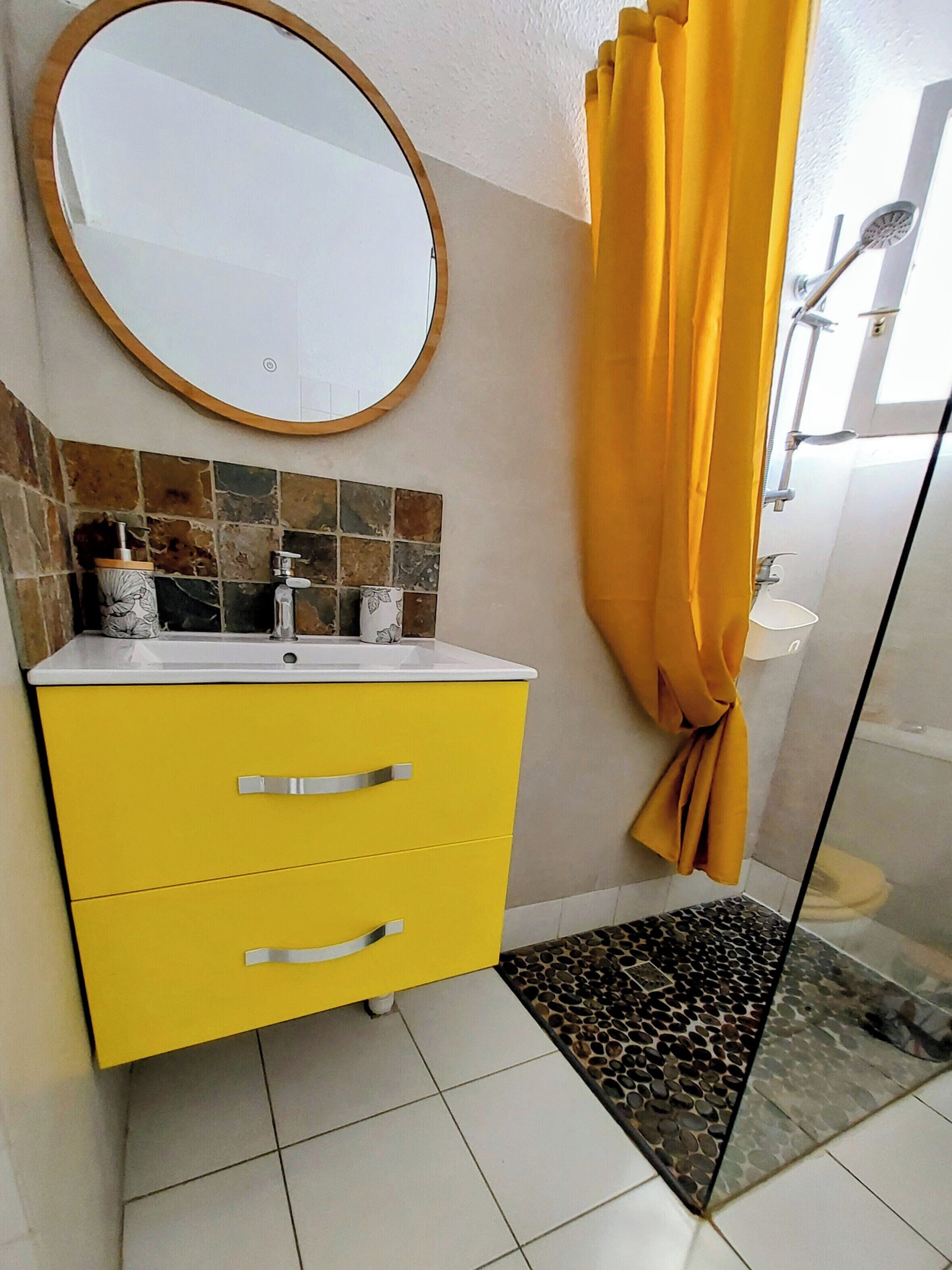 A bathroom with a yellow vanity, a round mirror, a shower with a glass door, and a tiled floor.