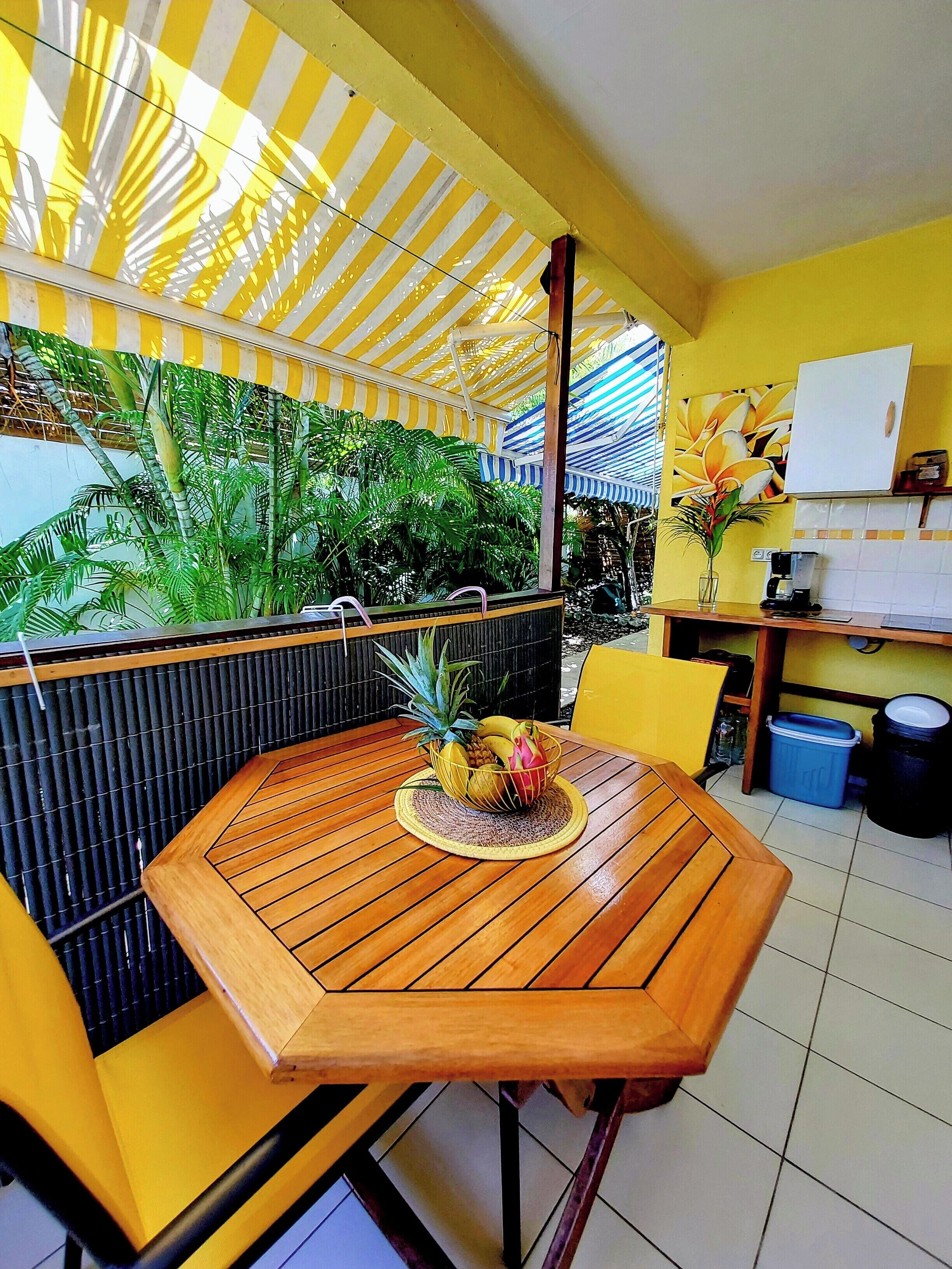 A wooden table with a woven placemat, a fruit bowl, and a yellow chair in a room with yellow walls and a striped ceiling.