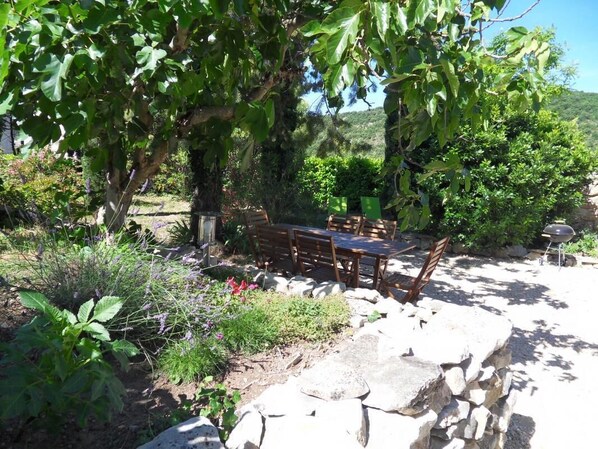 Un espace de restauration en plein air avec une table et des chaises en pierre, entouré d’une végétation luxuriante et offrant une vue sur les collines.