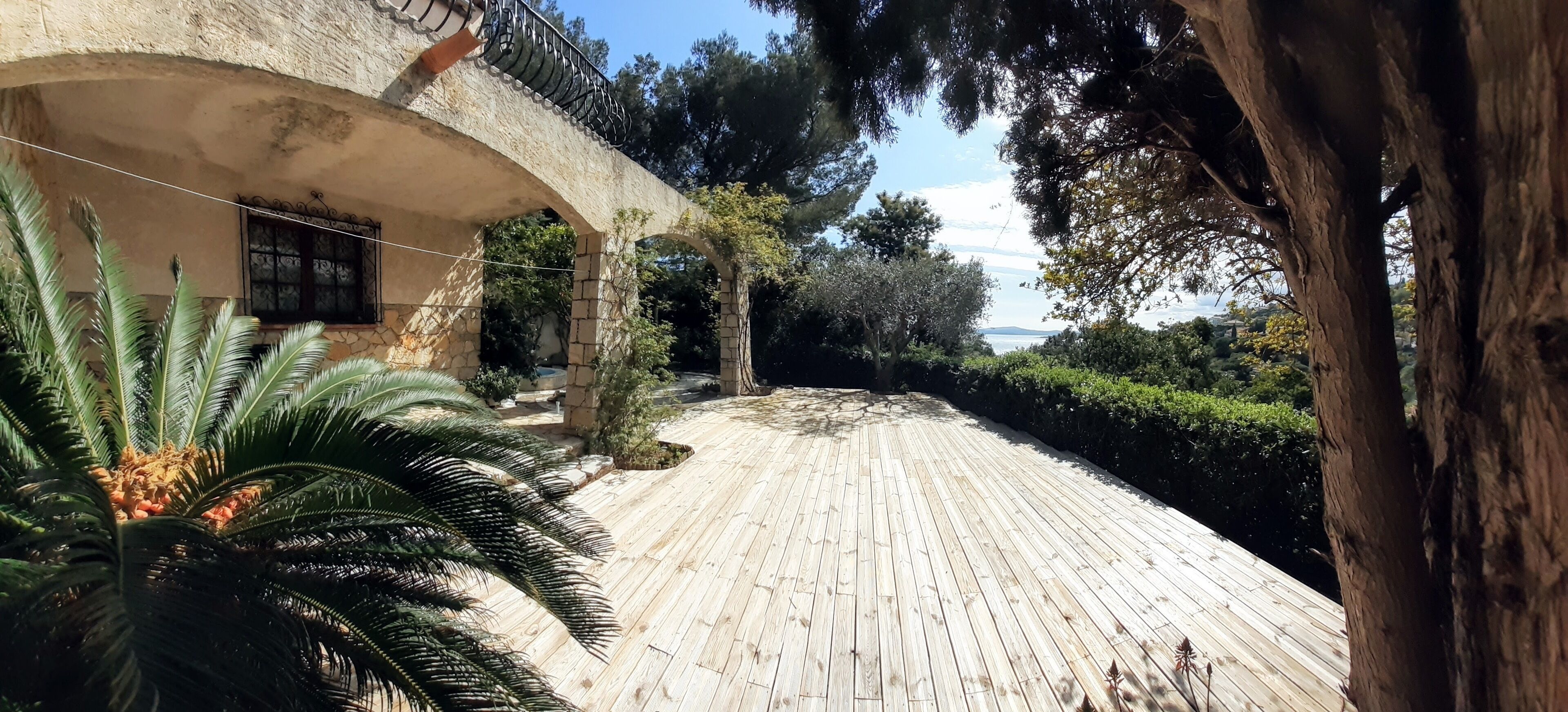 Una terraza de madera con un arco de piedra, palmeras y vista al mar.