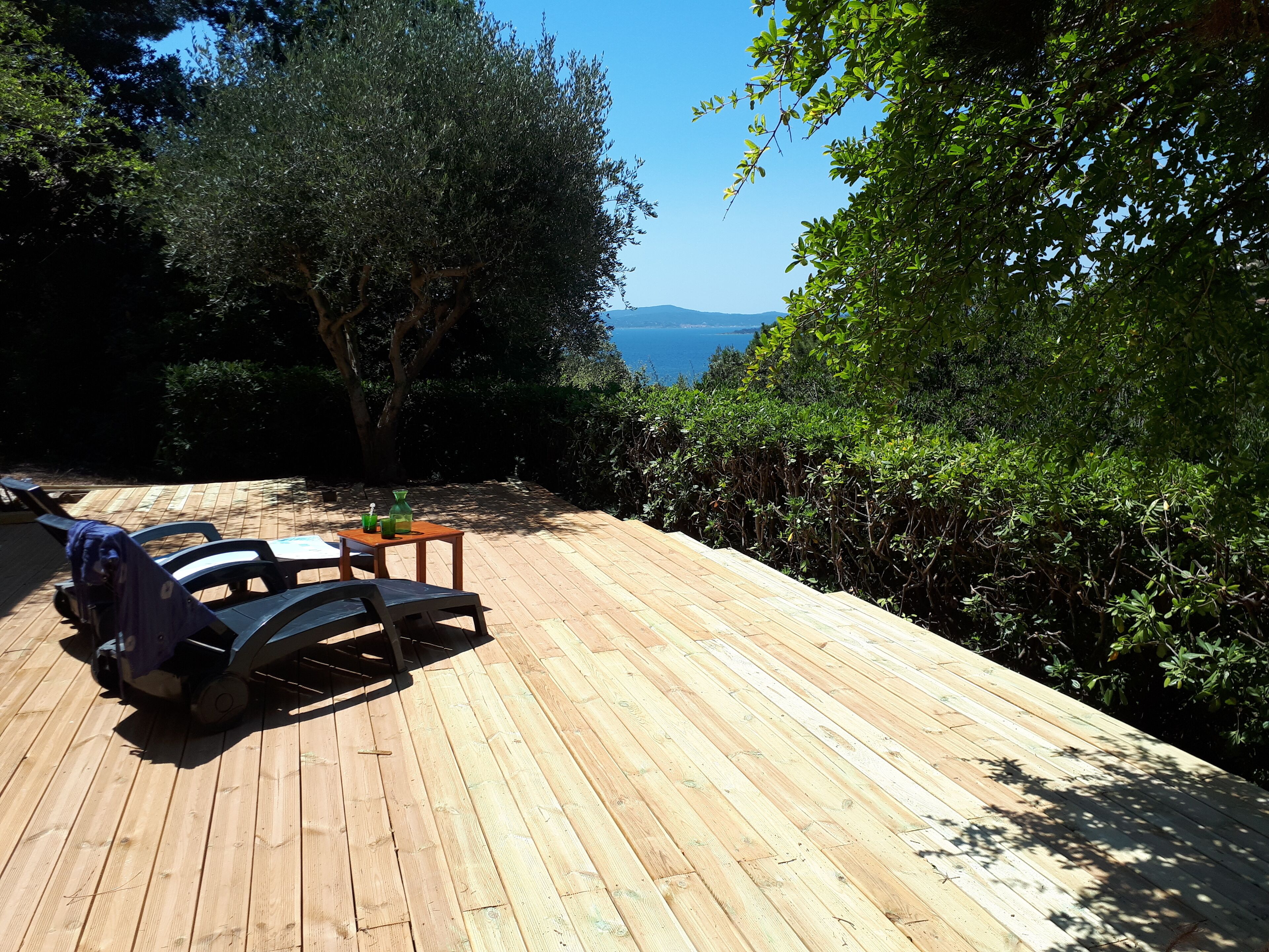 Eine Holzterrasse mit Gartenmöbeln, umgeben von üppigem Grün und Blick auf das Meer.
