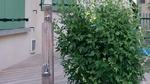 An outdoor shower with a wooden post, a green bush, and a building with green shutters in the background.