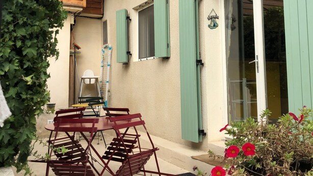 A wooden house with green shutters, a patio with a table and chairs, and a potted plant with red flowers.