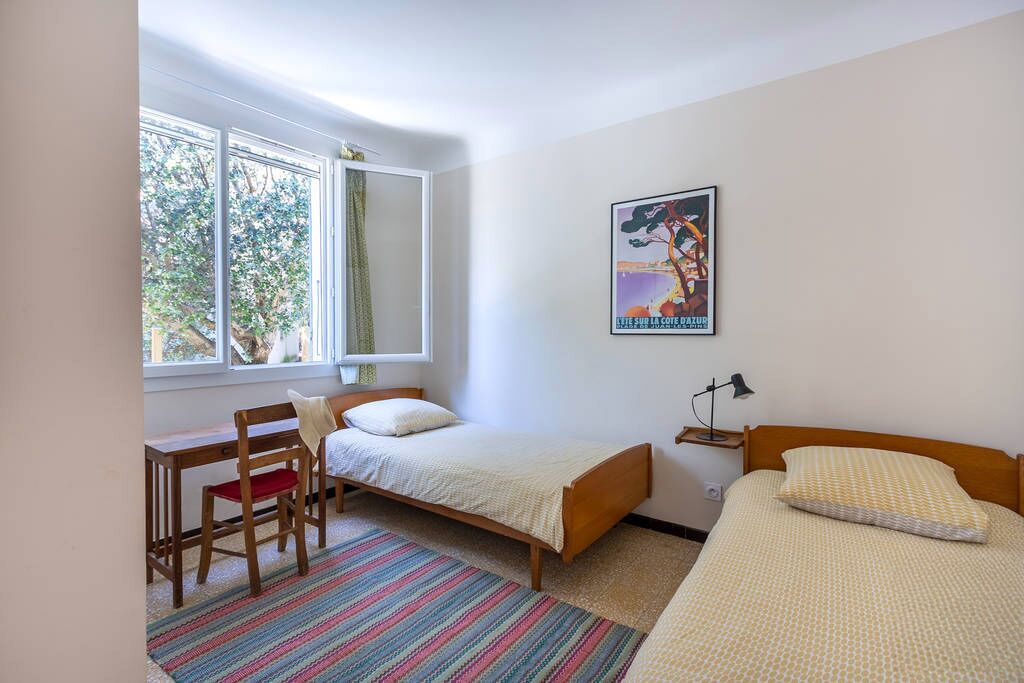 Ein Zimmer mit zwei Einzelbetten, einem Schreibtisch mit Stuhl, einem Fenster mit Blick auf Bäume und einem Poster an der Wand.