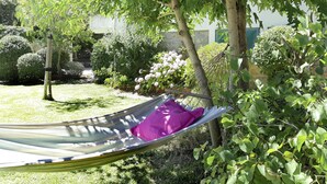 A hammock under a tree in a garden with various plants and a building in the background.