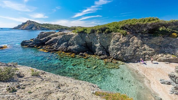 A rocky coastline with clear water, a sandy beach, and a hill with vegetation.