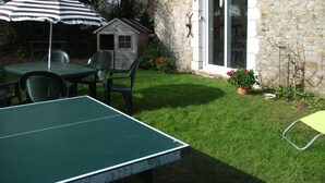 Una casita de madera, una mesa con sillas y una mesa de ping pong en un patio trasero con un muro de piedra y una puerta.