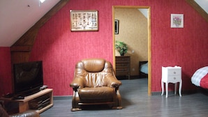 Una habitación con techo inclinado, un sillón de cuero marrón, un televisor sobre un mueble de madera, una mesita de noche blanca pequeña y una cama con manta de cuadros.
