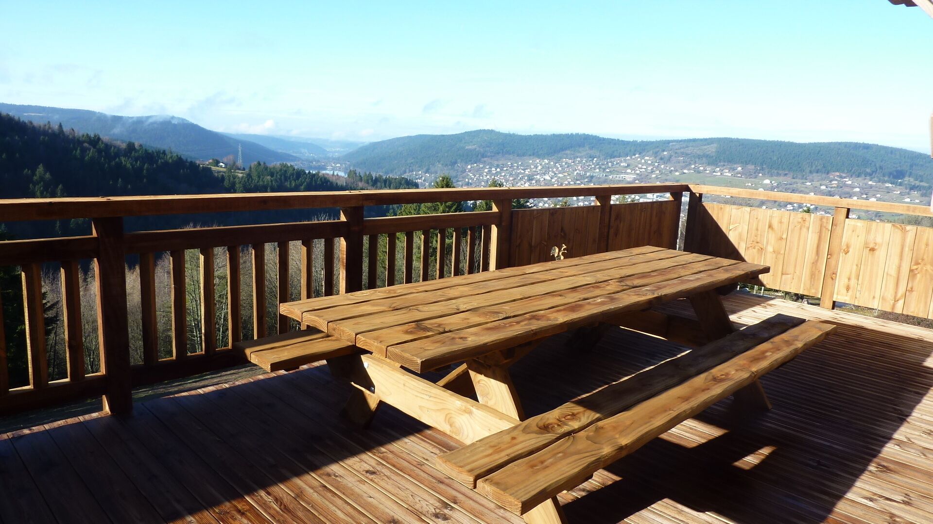 Una terraza de madera con una mesa de picnic y bancos, con vistas a un pueblo y montañas.