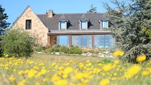 Una casa de piedra con techo inclinado, ventanas amplias y un jardín con flores amarillas.