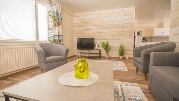 A living room with two grey armchairs, a coffee table with a yellow candle, a TV on a stand, and a kitchen in the background. - Jääskän Loma Kojola Apartments Alahärmä (Kauhava)