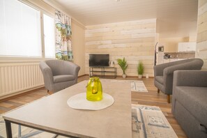 Una sala de estar con dos sillones grises, una mesa de centro con una vela amarilla, un televisor sobre un mueble bajo y una cocina al fondo.