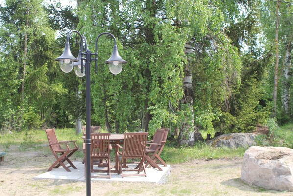 An outdoor dining area with a table and chairs, surrounded by trees and a large rock.