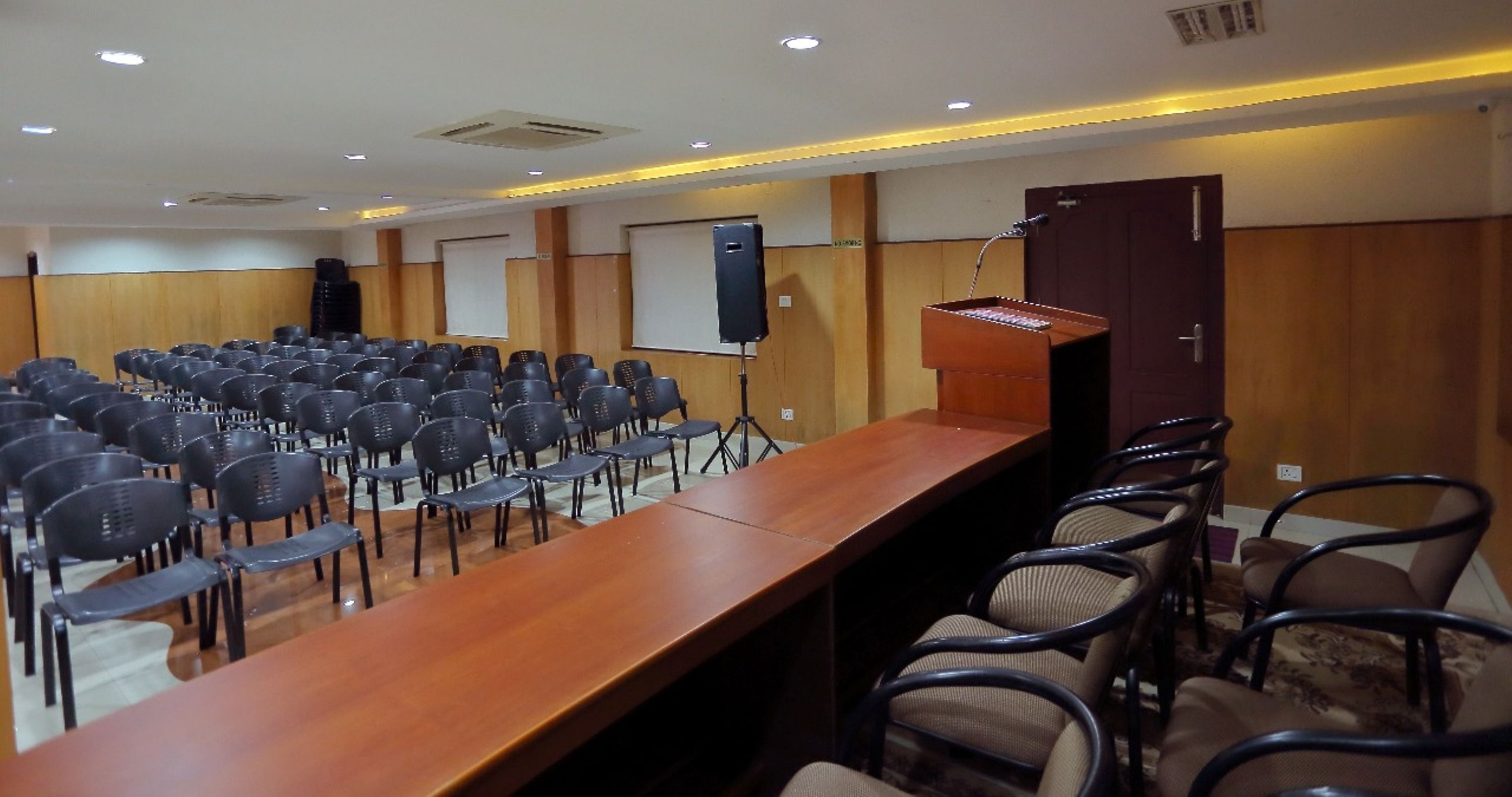 Un salón de conferencias con una larga mesa de madera, filas de sillas, un atril y una pantalla de proyector.