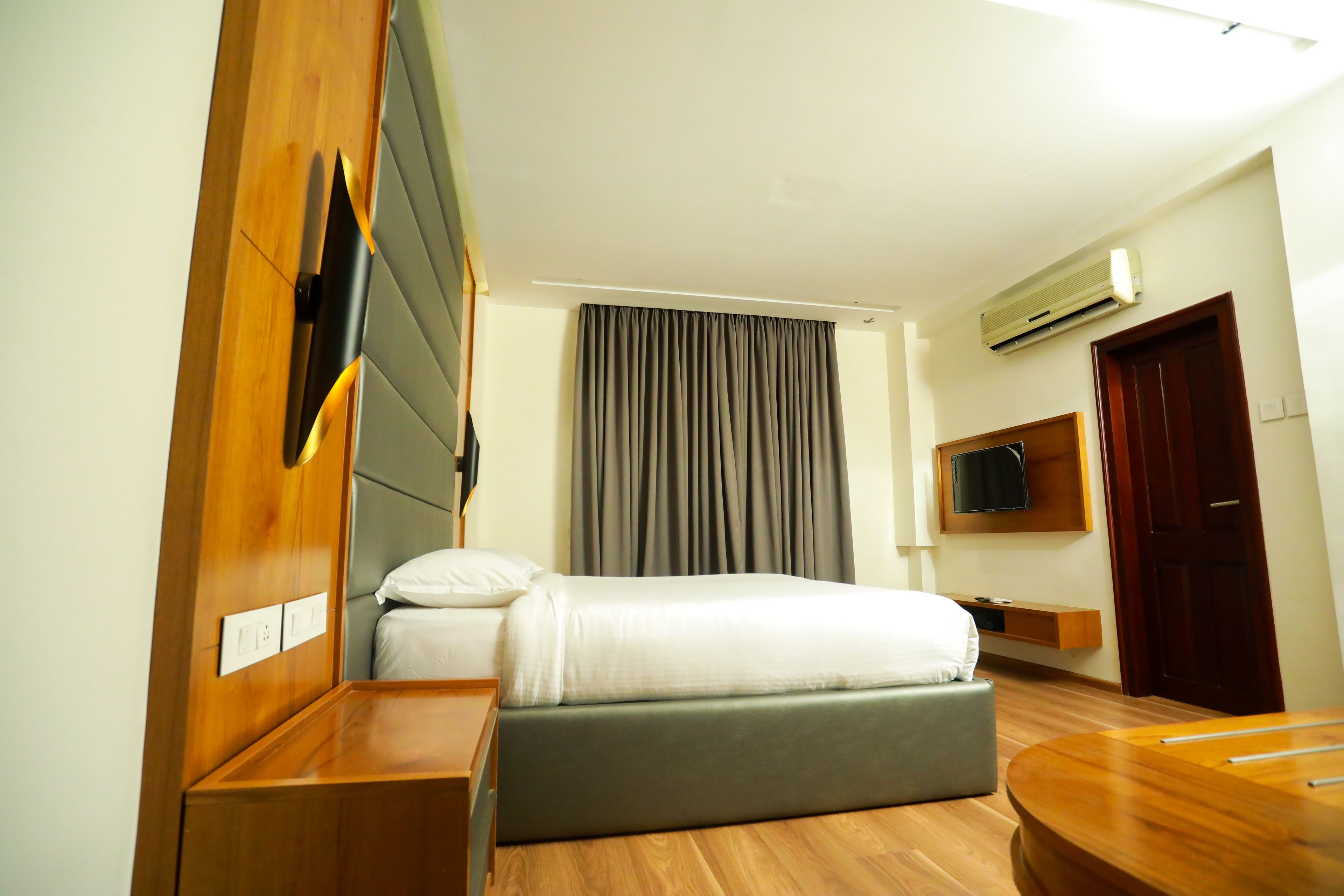 Ein modernes Hotelzimmer mit einem Holzkopfteil, einem Bett mit grauer Bettwäsche, einem Nachttisch aus Holz, einem kleinen Beistelltisch, einem fest installierten Fernseher und einer Klimaanlage.