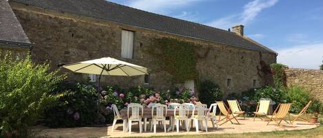 En bygning i sten med en havepatio, der inkluderer et bord, stole, en parasol og krukker med planter.