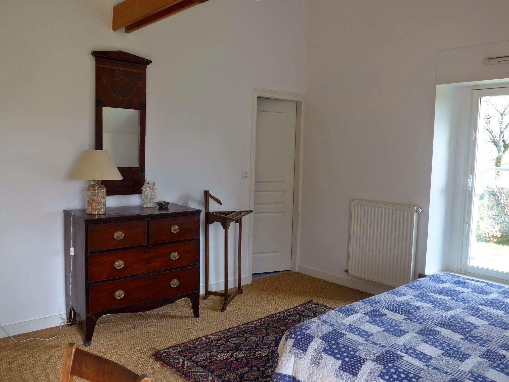 A bedroom with a wooden dresser, a mirror, a lamp, a chair, a bed with a blue quilt, a radiator, and a door.