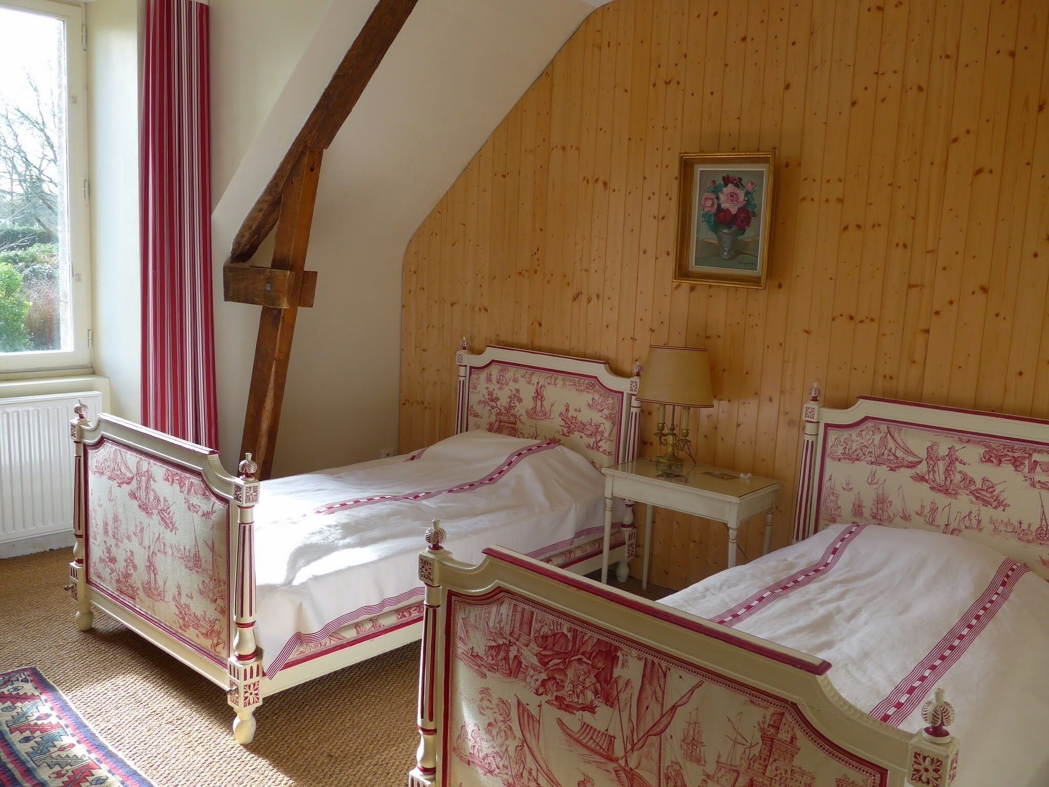 Ein Schlafzimmer mit zwei Betten, Holzwänden, einem Fenster mit roten Vorhängen und einem kleinen Nachttisch mit einer Lampe.