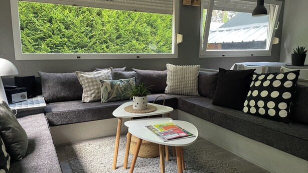 Ein modernes Wohnzimmer mit einem grauen Sofa, einem Couchtisch und Blick ins Grüne durch große Fenster.