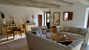 A living room with a sofa, armchairs, a coffee table, and a dining table with chairs. There is a wooden cabinet, a mirror, and a vase with branches.