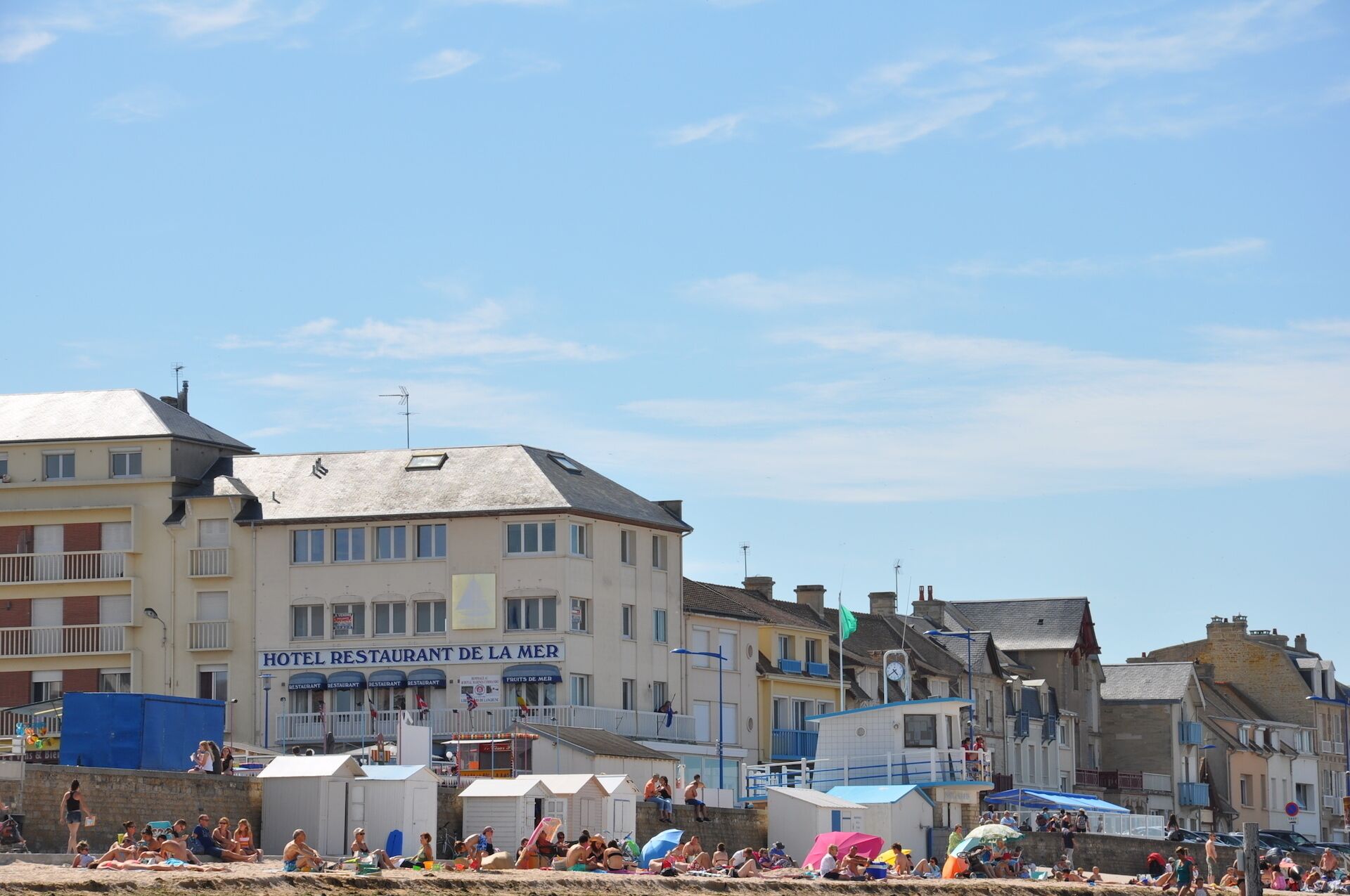 Ein mehrstöckiges Gebäude an der Küste, mit einem Restaurant namens 'HOTEL RESTAURANT DE LA MER' im Erdgeschoss und einem Strandabschnitt mit Personen und Sonnenschirmen.
