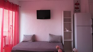 Une chambre à coucher avec un lit, un téléviseur fixé au mur, un réfrigérateur blanc et une fenêtre avec des rideaux rouges.