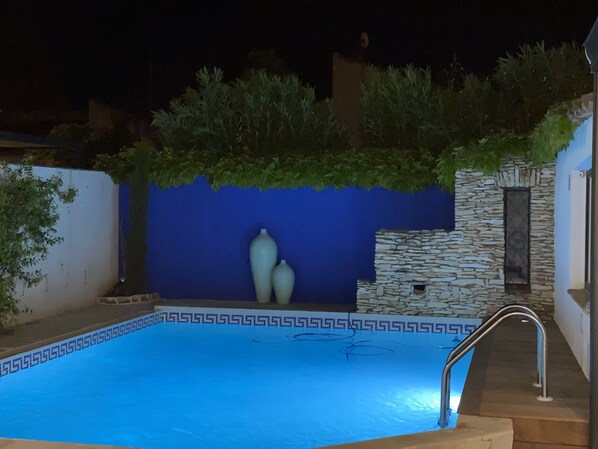A swimming pool with a blue mosaic border, a stone wall, and a small stone building with a window.