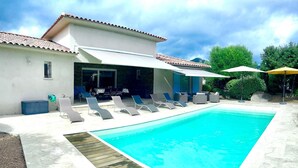 Ein Schwimmbecken mit Liegestühlen und Sonnenschirmen, eine überdachte Terrasse und ein Haus mit Ziegeldach.