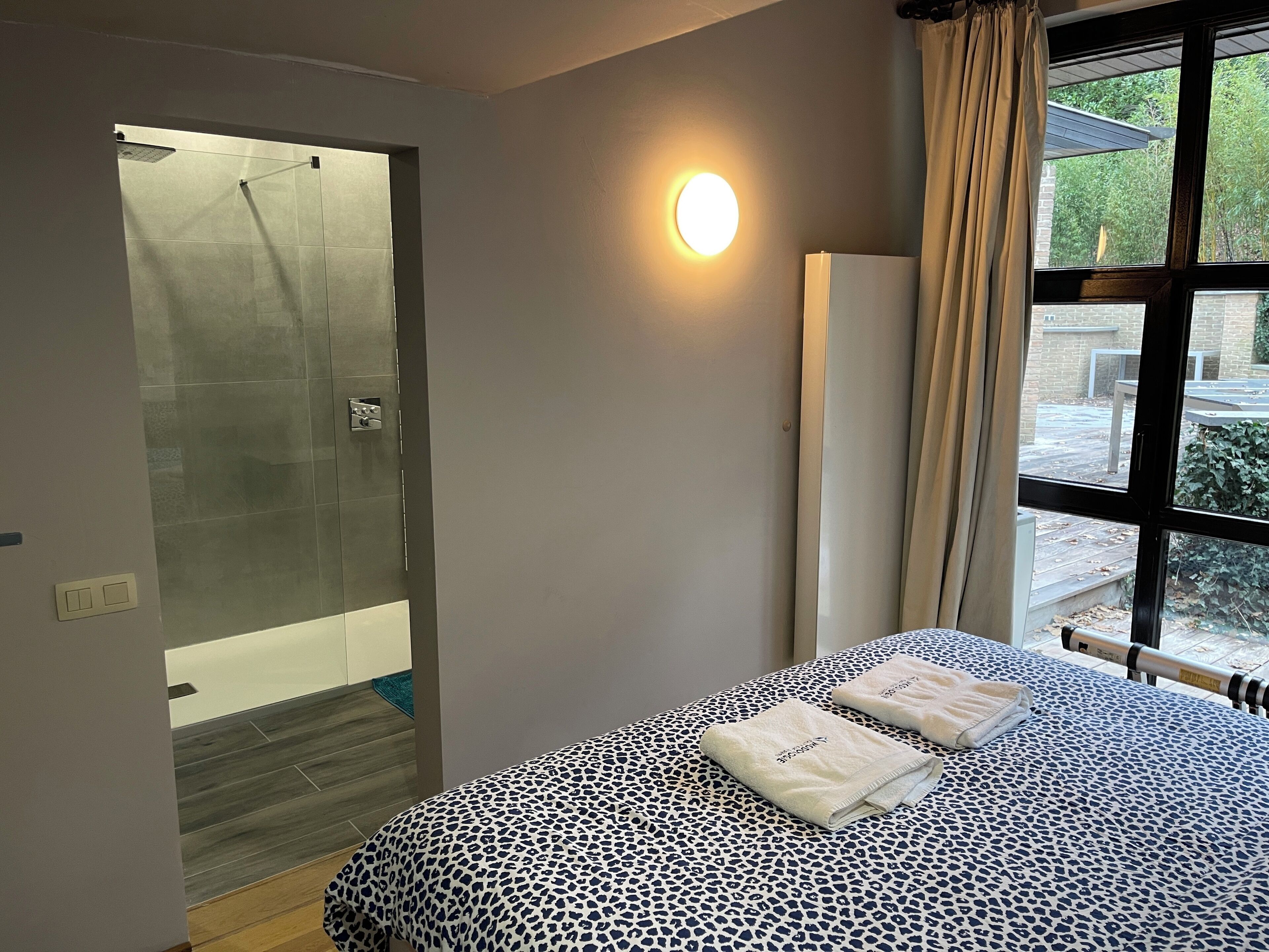 Une chambre d’hôtel avec une porte de douche en verre, un lit avec une literie à motifs léopard et une lampe de chevet.