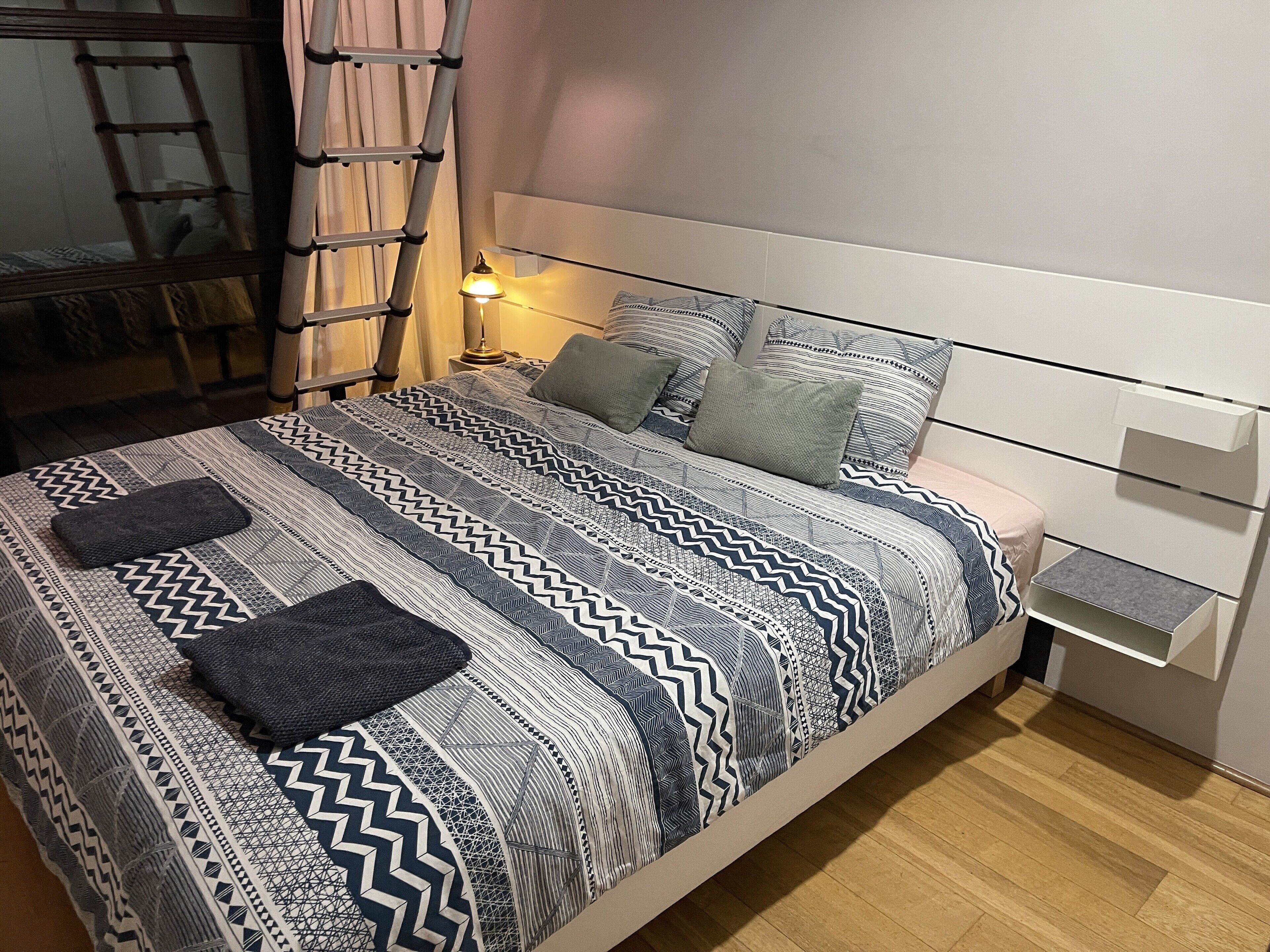 A neatly made bed with patterned bedding, a bedside table with a lamp, and a wooden ladder against the wall.